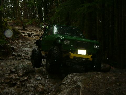 1990 Ford Bronco - Off-road truck driving through a challenging, muddy, and rocky path in a dense forest.