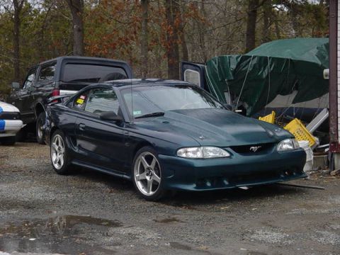 1995 Ford Mustang - A dark green Ford Mustang is parked outside next to other vehicles and covered items.