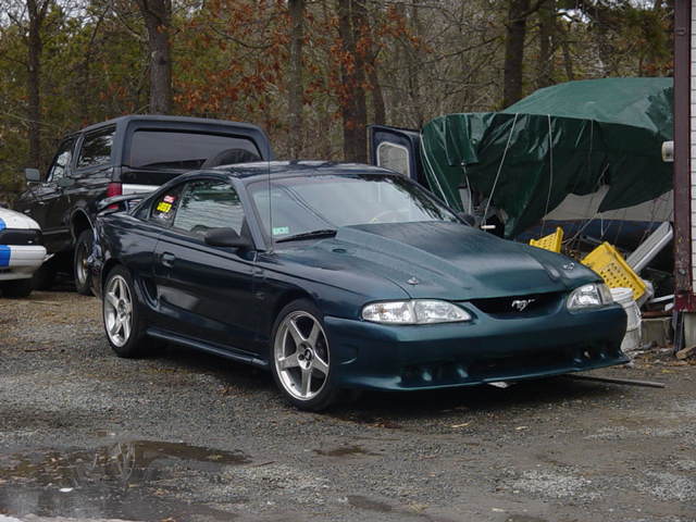 Dark green Ford Mustang with a hood scoop, parked on gravel. A black SUV is behind it