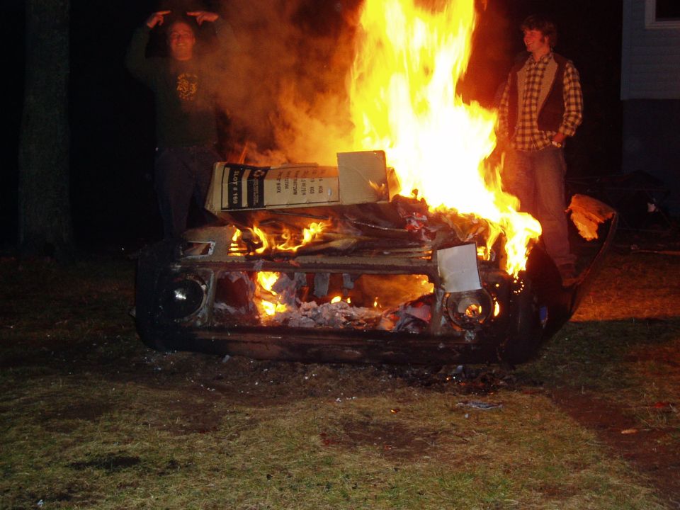 Car engulfed in flames, two people standing nearby. Cardboard boxes are on top of the burning car