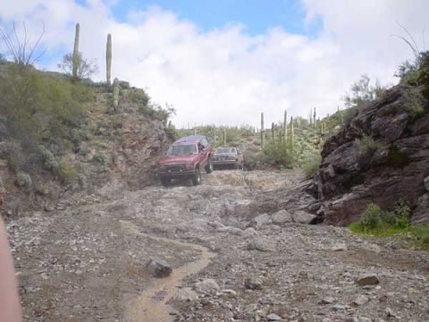1989 Ford Bronco - A red 1989 Ford Bronco navigates a desert path, with another vehicle visible in the distance.