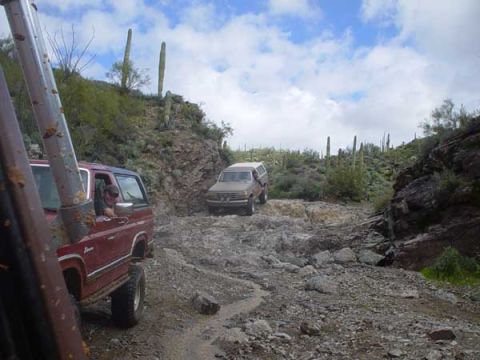 1989 Ford Bronco - Two SUVs navigate a challenging rocky path through a desert landscape under a cloudy sky.