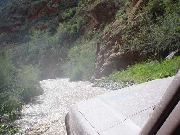 Vehicle driving through a shallow, rocky river in a canyon. Part of the vehicle's hood and windshield frame are visible