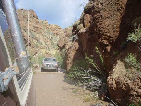 1989 Ford Bronco - A silver Ford Bronco navigates a water-filled path between rocky canyon walls.