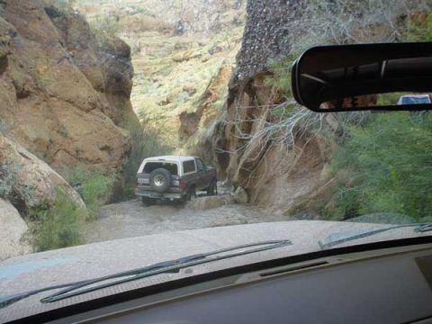 1989 Ford Bronco - A 1989 Ford Bronco navigates a narrow, water-filled canyon, its rear visible as it drives through the rocky terrain.