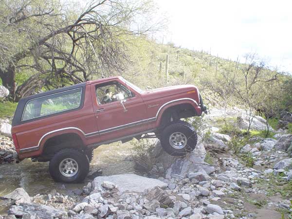 Red Ford Bronco with black top driving over rocks in a stream, desert landscape in background