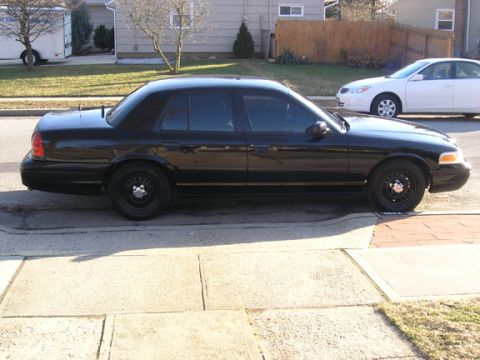1999 Ford Crown Victoria - A black sedan is parked on the street next to a sidewalk, with a white car visible in the background.