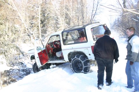 2003 Jeep TJ - A white SUV with snow chains on its tires is stuck in deep snow off a wooded path.