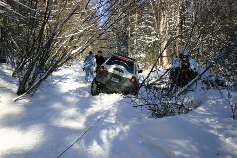 2003 Jeep TJ - Two people assist a white and black SUV that is stuck in a snowy wooded area, with a tow rope attached.
