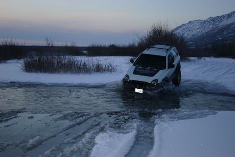 2003 Jeep TJ - A white SUV navigates through a snowy landscape with water and ice, under a dusky sky with mountains in the distance.