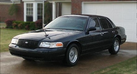 1999 Ford Crown Victoria - A black sedan is parked in a driveway in front of a house and garage. The vehicle has chrome wheels.