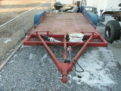 1995 Ford Bronco - A dual-axle flatbed utility trailer with a red-painted steel frame and diamond-plate deck sits on a snowy gravel lot beside a chain-link fe…