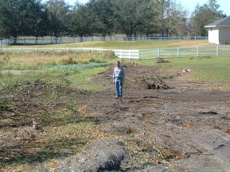 Man standing in cleared field with debris and white fence in background