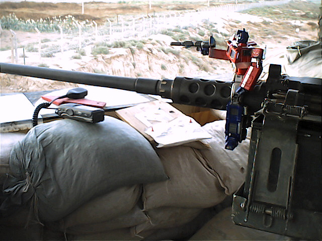 Optimus Prime toy standing on a large machine gun barrel, with sandbags and a radio in the foreground