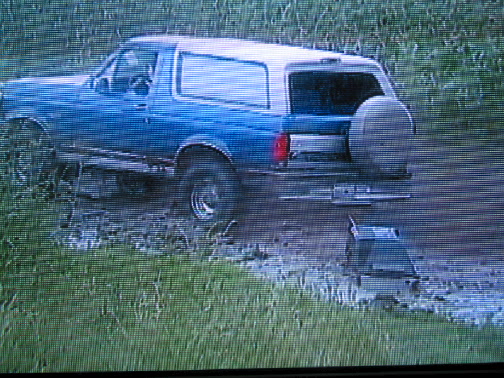 Blue and white Ford Bronco SUV stuck in mud on a grassy trail, rear three-quarter view