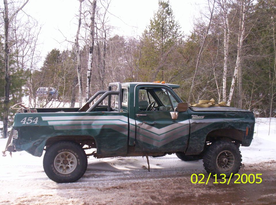 Green pickup truck with a roll bar in the bed, lifted suspension, and custom side graphics. Another vehicle is visible in the background