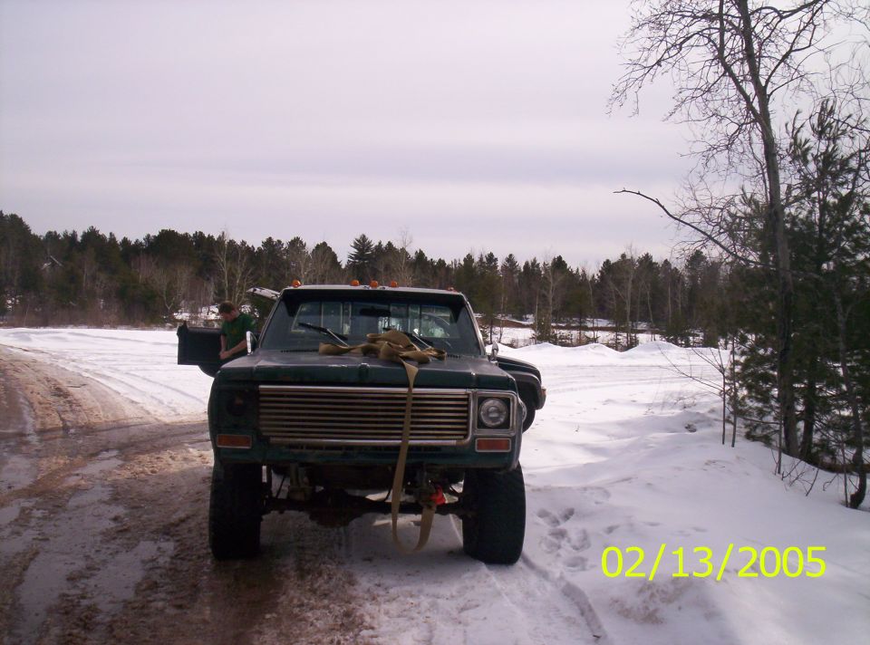 Dark green pickup truck with a chrome grille and straps across the hood, parked on a snowy road