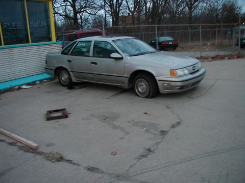 1997 Ford Taurus - A silver sedan sits parked on cracked asphalt in front of a building with a flea market sign.