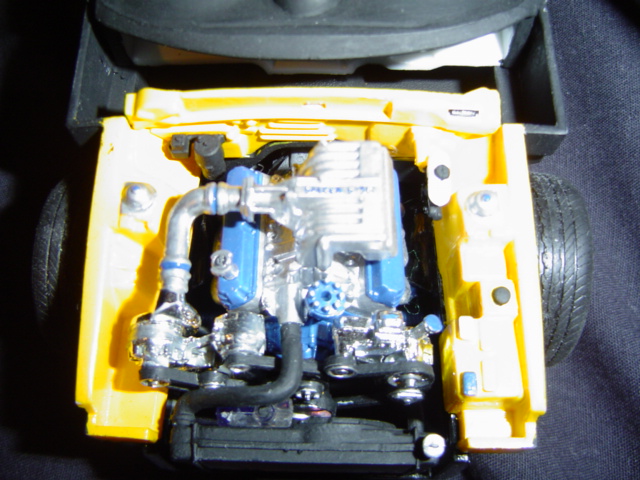 Yellow model car engine bay with a silver and blue engine, chrome hoses, and black tires visible