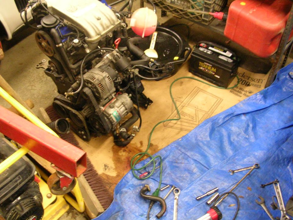 Engine sitting on a garage floor mid-swap beside a tarp of wrenches and a battery