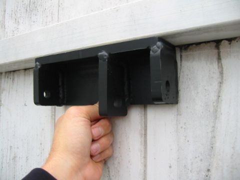 1992 Ford Bronco - A hand holds up a shop-fabricated black-painted track bar bracket with visible weld beads, a center double-shear tab and side mounting hole…