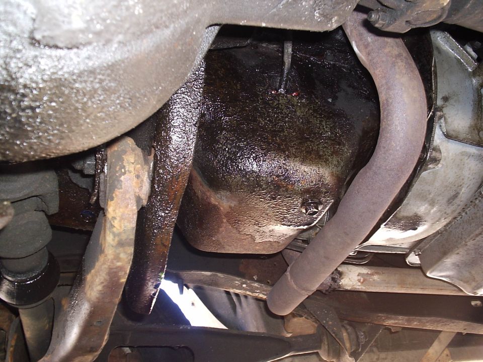 Underside view of an oil-soaked engine oil pan with rusty exhaust pipes and frame rail