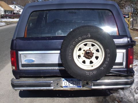 1985 Ford Bronco - Rear view of a dark blue Ford Bronco with a swing-away tire carrier holding a Firestone ATX radial on a white eight-slot steel wheel. The t…