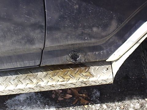 1985 Ford Bronco - Close side view of a dark blue truck's lower cab corner and rocker panel showing a rusted-through hole in the sheet metal above a diamond-p…