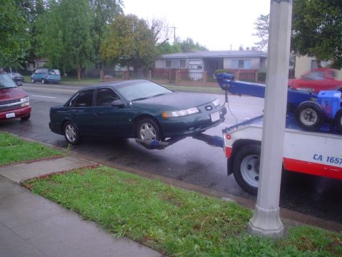1993 Ford Taurus - A green sedan is towed by a truck on a wet street, with other vehicles visible in the background.