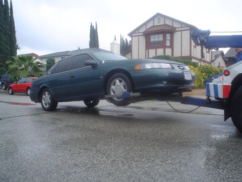 1993 Ford Taurus - Green 1993 Ford Taurus is loaded onto a tow truck with other vehicles visible on the street.