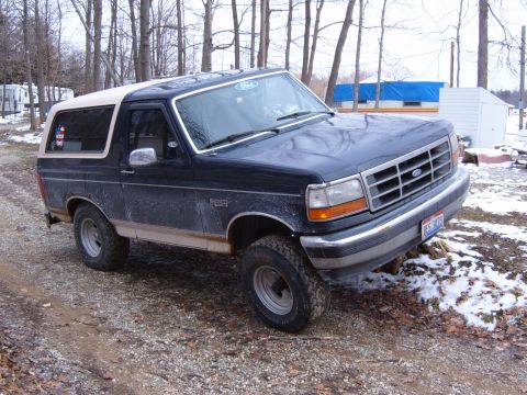 1993 Ford Bronco - A dark blue Ford Bronco with a tan roof sits on a gravel surface, with trees and buildings in the background.