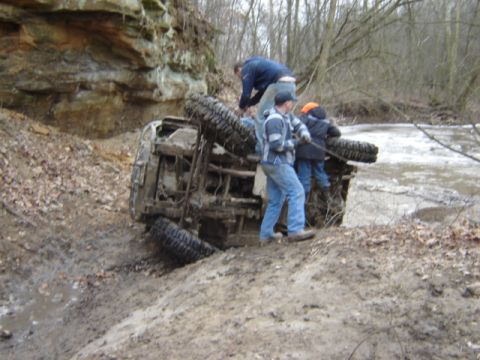 1992 Ford Bronco - People attempt to recover an overturned off-road vehicle stuck in mud near a creek.