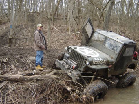 1992 Ford Bronco - A man watches as a vintage military jeep gets stuck in the mud and water in a wooded area.