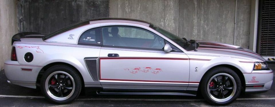 Silver Mustang coupe side view with red stripes, Cobra decals, black wheels and red calipers by a concrete wall