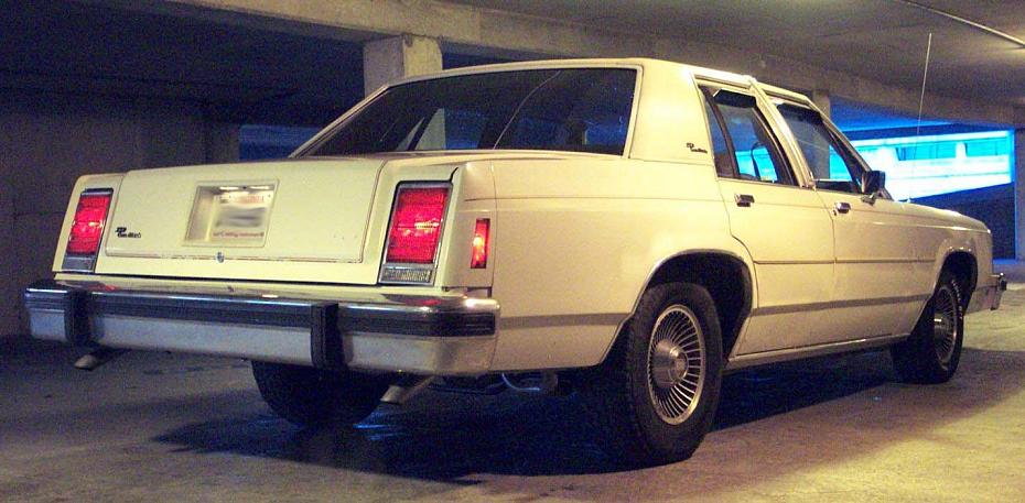Rear view of a cream-colored 1983 Ford sedan parked indoors. It has rectangular taillights and chrome bumpers