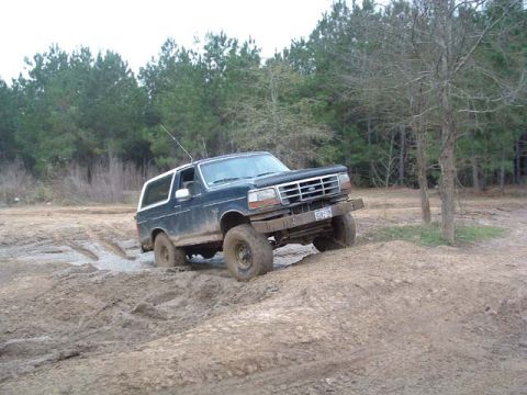 1993 Ford Bronco - This Ford Bronco navigates a challenging muddy terrain with trees in the background.