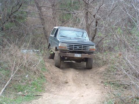 1993 Ford Bronco - A dark gray Ford Bronco navigates a wooded trail with a lifted suspension and large tires.