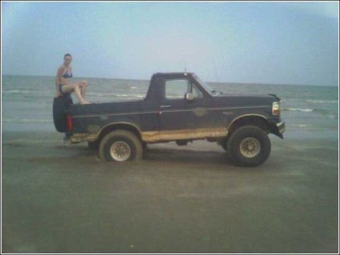 1993 Ford Bronco - A woman sits on the spare tire of a 1993 Ford Bronco partially submerged in sand and water at the beach.