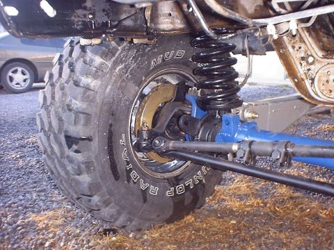 1988 Ford Bronco - A close-up view of the suspension and a large off-road tire on a vehicle. Another car is visible in the background.