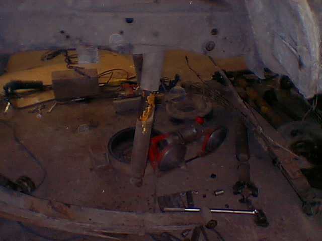 Dim overhead shot of a workbench littered with tools, parts and a red component under a vehicle body