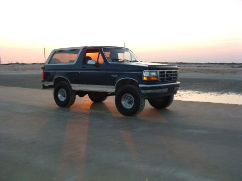 1993 Ford Bronco - A dark blue 1993 Ford Bronco with a tan top is parked on a sandy beach during sunset.