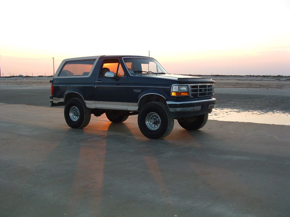 Dark blue lifted Ford Bronco parked on beach sand at sunset, front three-quarter view