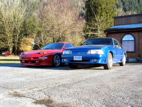 1994 Ford Probe - A red Ford Probe and a blue Ford Mustang are parked in front of a wooden building.