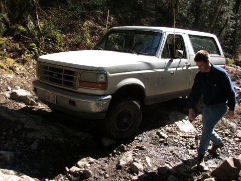 1980 Ford Bronco - White Ford Bronco on a rough, wooded trail. A man walks alongside the vehicle.