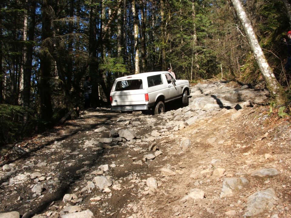 White SUV navigating a very rocky, wooded trail. The vehicle is seen from the rear, going uphill
