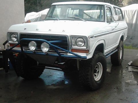 1978 Ford Bronco - Freshly painted white full-size Ford Bronco on wet pavement, shot from the front three-quarter angle with a blue tube prerunner bumper, two…