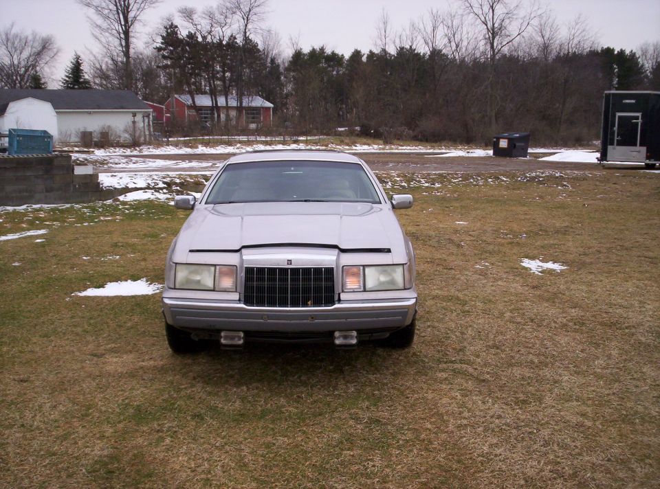 Front view of a silver sedan parked on grass with some snow patches. Buildings and trees in the background
