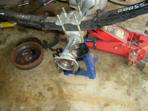 1995 Ford Bronco - A disassembled vehicle axle and leaf spring assembly rests on a garage floor with a red hydraulic jack and blue jack stands.