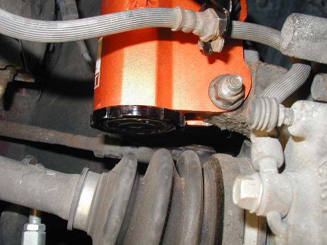 Close-up of an orange automotive part, possibly a reservoir, with brake lines and suspension components visible