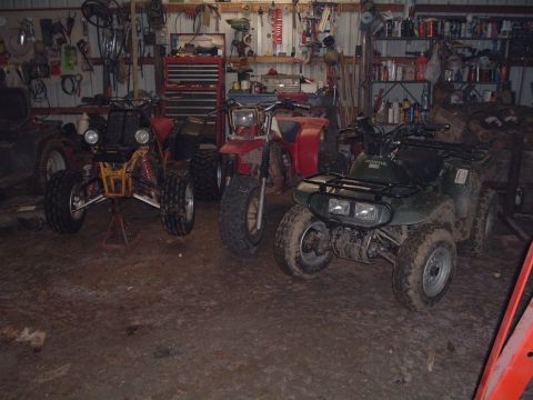 1995 Mercury Grand Marquis - Three ATVs are parked in a cluttered garage, with tools and supplies visible on shelves and walls.